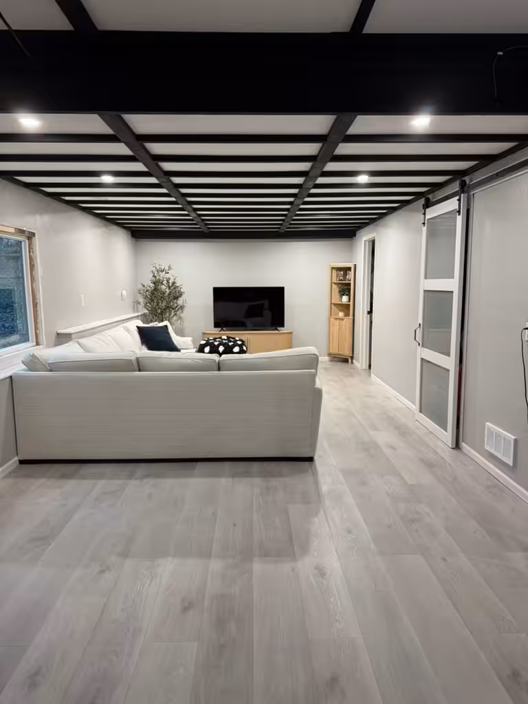 Modern living room with light wood flooring, white sectional sofa, TV on a wooden stand, a corner plant, wooden bookshelf, and black ceiling beams. Sliding glass door on the right. Room is well-lit and spacious.