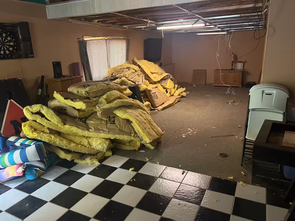 A basement room with checkered flooring and carpet, exposed ceiling, piles of yellow insulation on the floor, scattered debris, and furniture pushed to the sides. The space appears to be under renovation or repair.