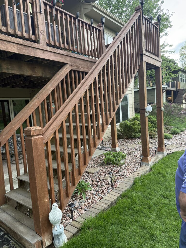 Wooden outdoor staircase with railing leads from an upper deck to a landscaped yard with rocks and plants. Part of a house, lawn, and a person in a blue shirt are also visible.